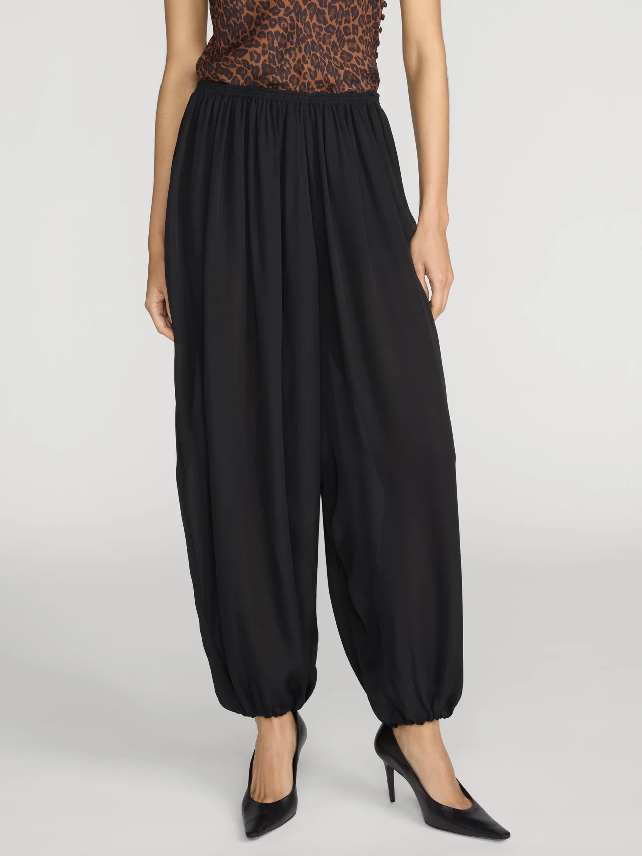 A model wearing black harem-style pants with an elastic waist and gathered cuffs, paired with a leopard-print top and black heels, against a light gray studio background.