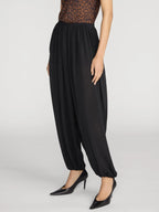 A model wearing black high-waisted, loose-fitting harem-style pants with gathered cuffs, paired with a leopard-print top and black pointed-toe heels, against a light gray studio background.