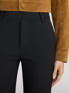 Close-up of a person wearing black tailored trousers and a tan suede cropped jacket, with one hand visible against a light gray studio background.