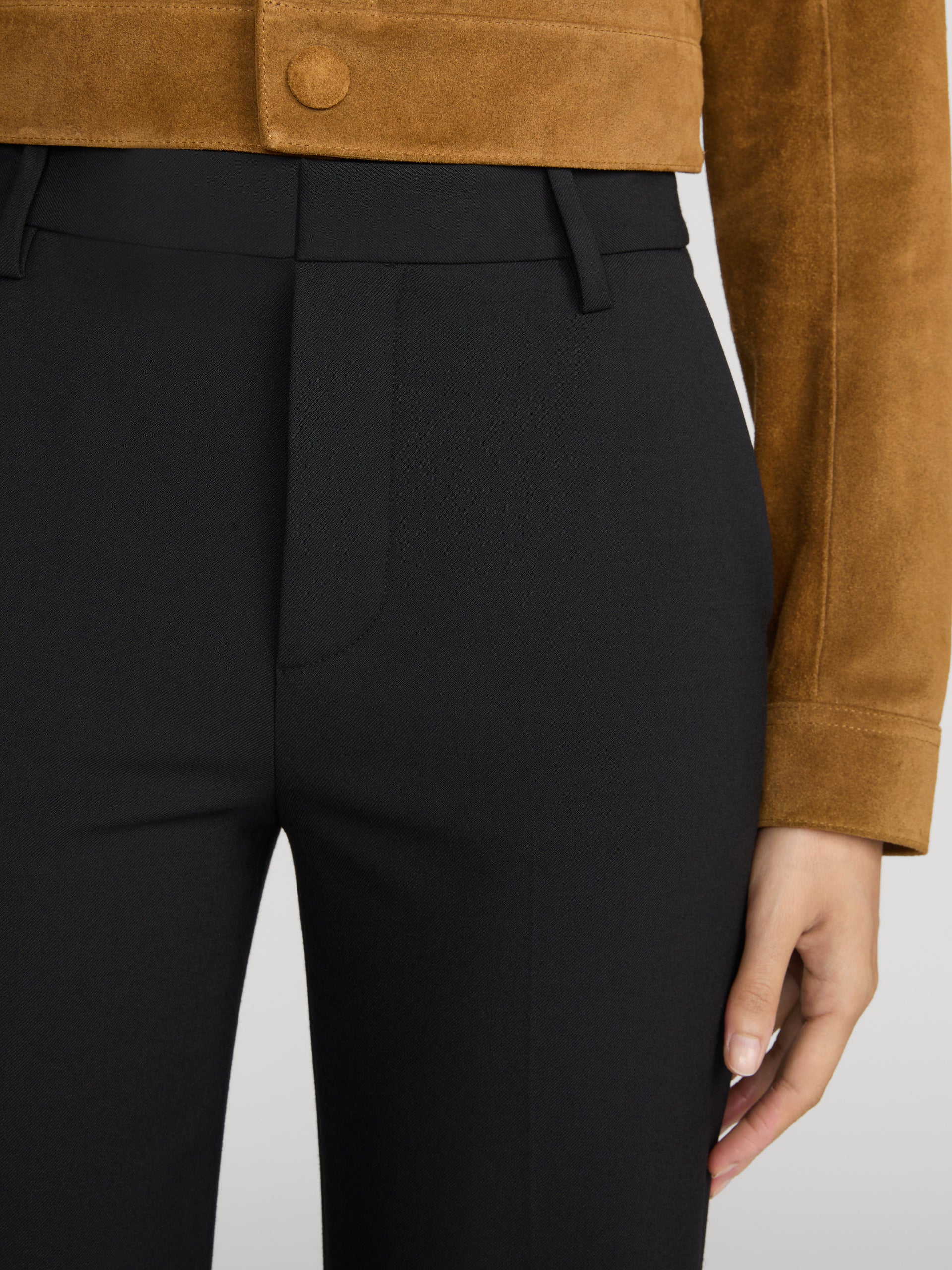 Close-up of a person wearing black tailored trousers and a tan suede cropped jacket, with one hand visible against a light gray studio background.
