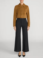 A model is shown from the neck down wearing a cropped tan suede jacket and black wide-leg trousers against a light gray studio background.