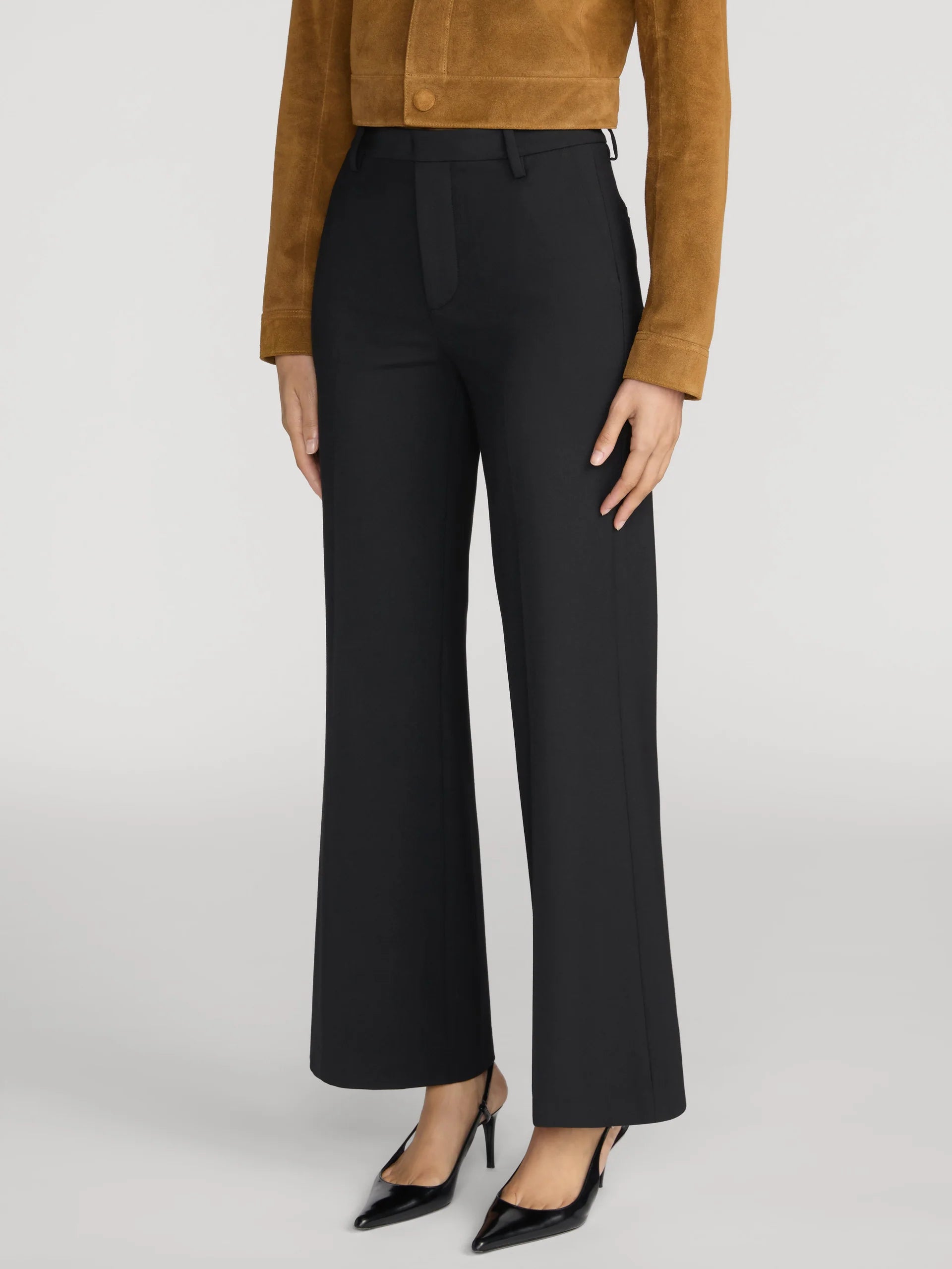 A model is shown from the waist down wearing black wide-leg trousers, a brown suede cropped jacket, and black pointed-toe heels against a light gray studio background.