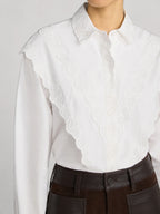 A close-up of a white long-sleeve blouse with a pointed collar and ornate scalloped eyelet lace trim down the front, worn with dark brown trousers against a light gray studio background.