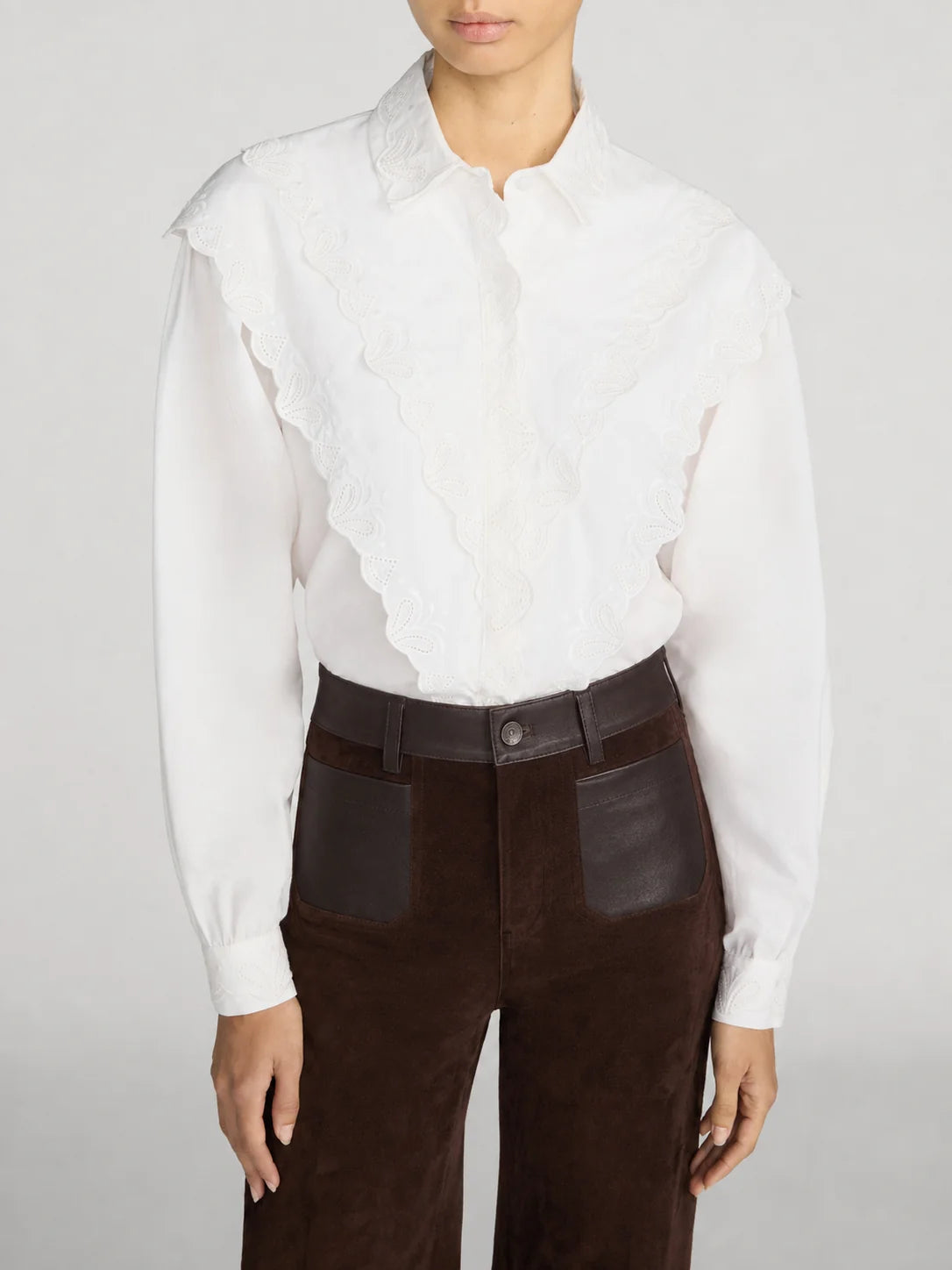 A model wearing a white long-sleeve blouse with a ruffled lace overlay across the chest and dark brown high-waisted trousers with leather patch pockets, against a light gray studio background.