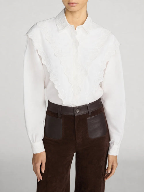 A model wearing a white long-sleeve blouse with a ruffled lace overlay across the chest and dark brown high-waisted trousers with leather patch pockets, against a light gray studio background.