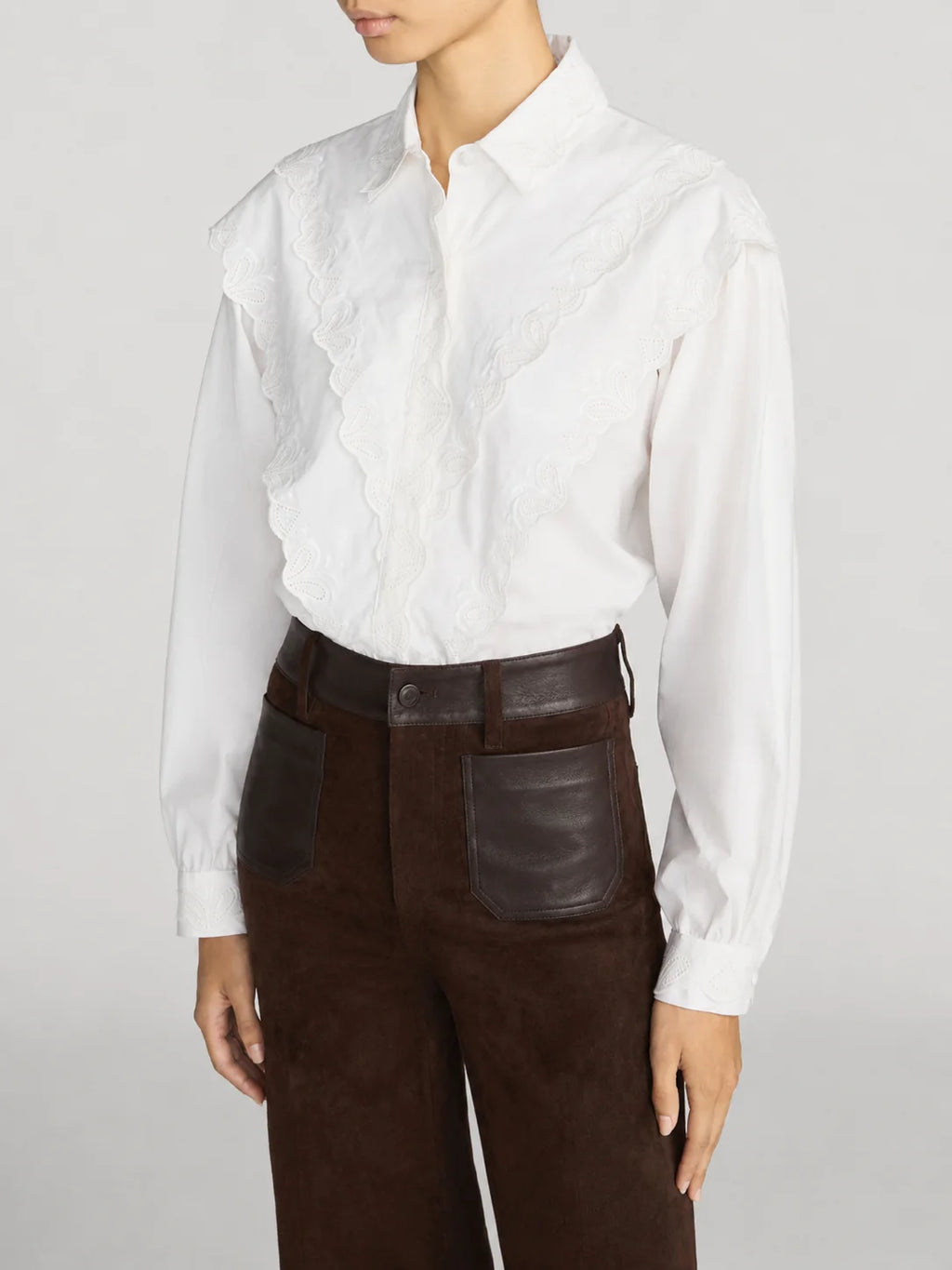 A model wearing a white long-sleeve blouse with a ruffled lace front panel and dark brown high-waisted trousers with leather patch pockets, against a light gray studio background.