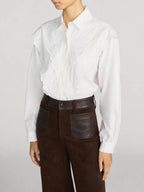 A model wearing a white long-sleeve blouse with a ruffled lace front panel and dark brown high-waisted trousers with leather patch pockets, against a light gray studio background.