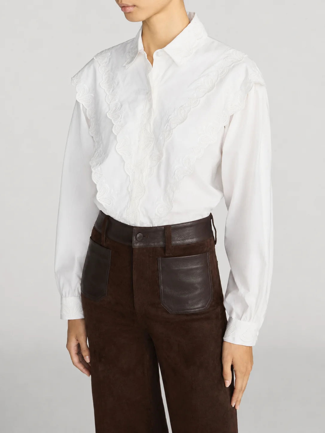 A model wearing a white long-sleeve blouse with a ruffled lace front panel and dark brown high-waisted trousers with leather patch pockets, against a light gray studio background.