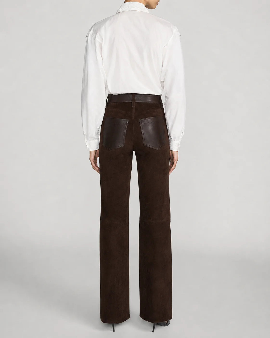 A model shown from the back wearing dark brown wide-leg trousers with dark brown leather patch pockets and a white button-down shirt, set against a light gray studio background.