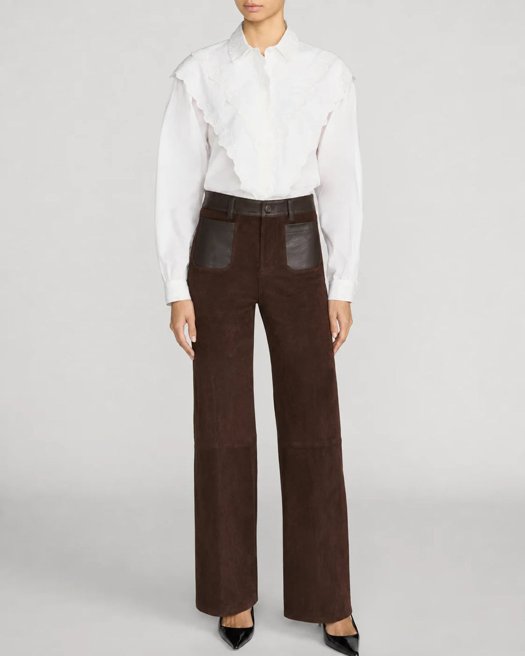 A model wearing high-waisted dark brown wide-leg trousers with dark brown leather patch pockets, styled with a white ruffled blouse and black pointed-toe heels against a light gray studio background.