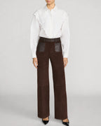 A model wearing high-waisted dark brown wide-leg trousers with dark brown leather patch pockets, styled with a white ruffled blouse and black pointed-toe heels against a light gray studio background.