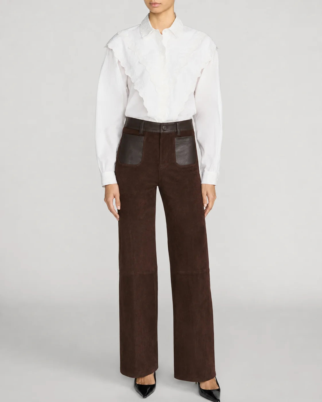 A model wearing high-waisted dark brown wide-leg trousers with dark brown leather patch pockets, styled with a white ruffled blouse and black pointed-toe heels against a light gray studio background.