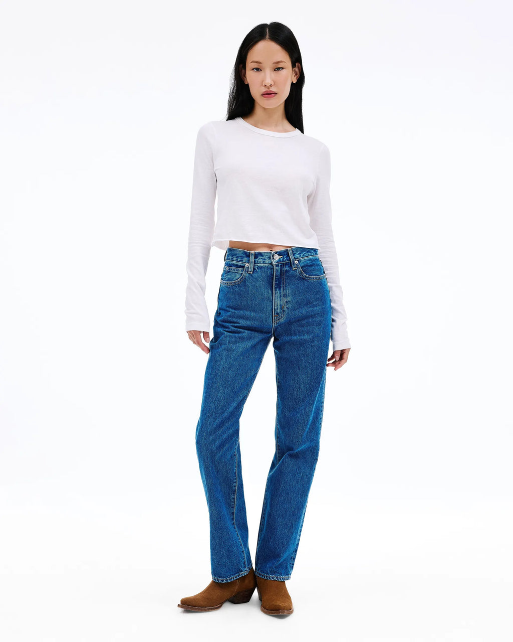A model stands against a white studio background wearing a white long-sleeve cropped top, blue high-waisted straight-leg jeans, and brown ankle boots.