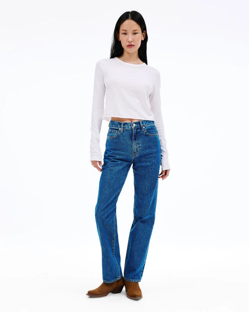 A model stands against a white studio background wearing a white long-sleeve cropped top, blue high-waisted straight-leg jeans, and brown ankle boots.