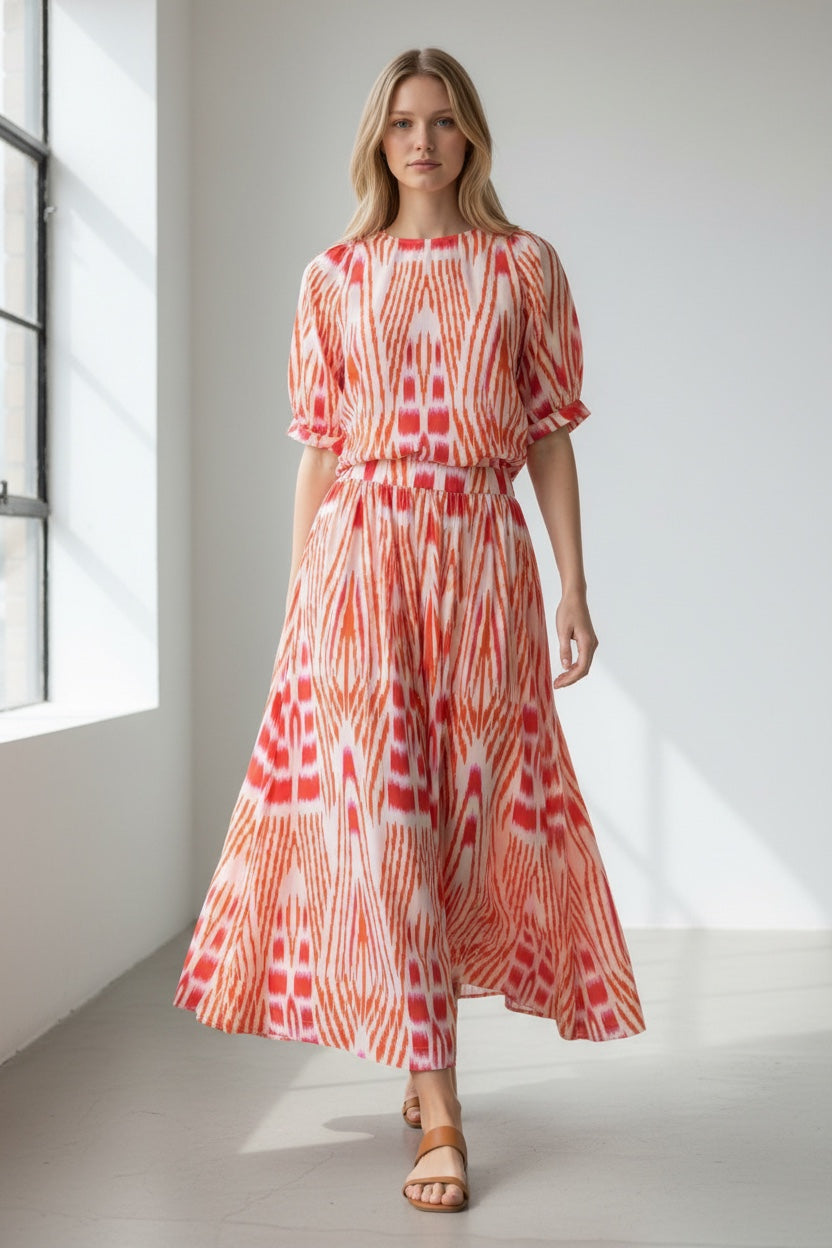 A woman modeling a short-sleeved midi dress with a red, orange, and white ikat pattern, standing in a bright, minimalist studio with a large window on the left.