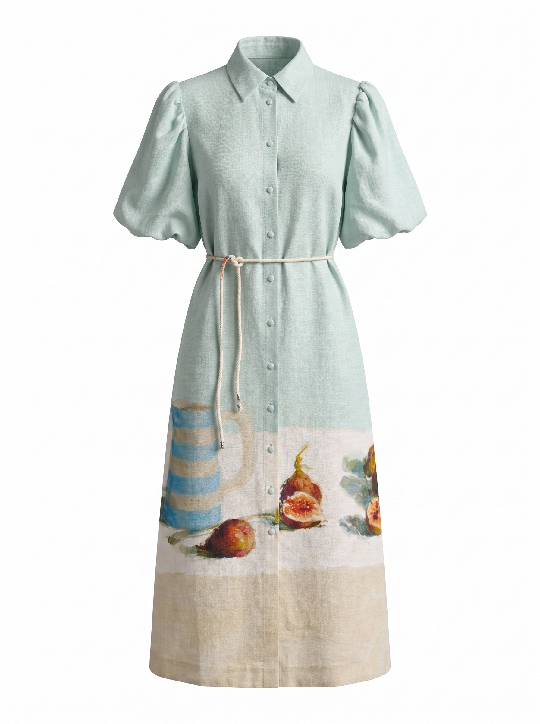 A light mint green linen shirt dress with puff sleeves, a button-down front, and a cream-colored printed border featuring a blue-and-white striped pitcher and figs at the hem.