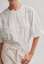 A close-up of a person wearing a white short-sleeve blouse with intricate floral embroidery and applique across the chest and shoulders, paired with light beige trousers.