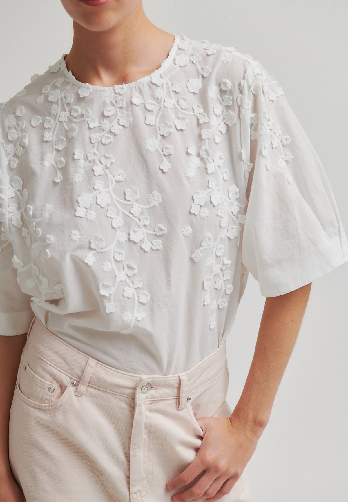 A close-up of a person wearing a white short-sleeve blouse with intricate floral embroidery and applique across the chest and shoulders, paired with light beige trousers.