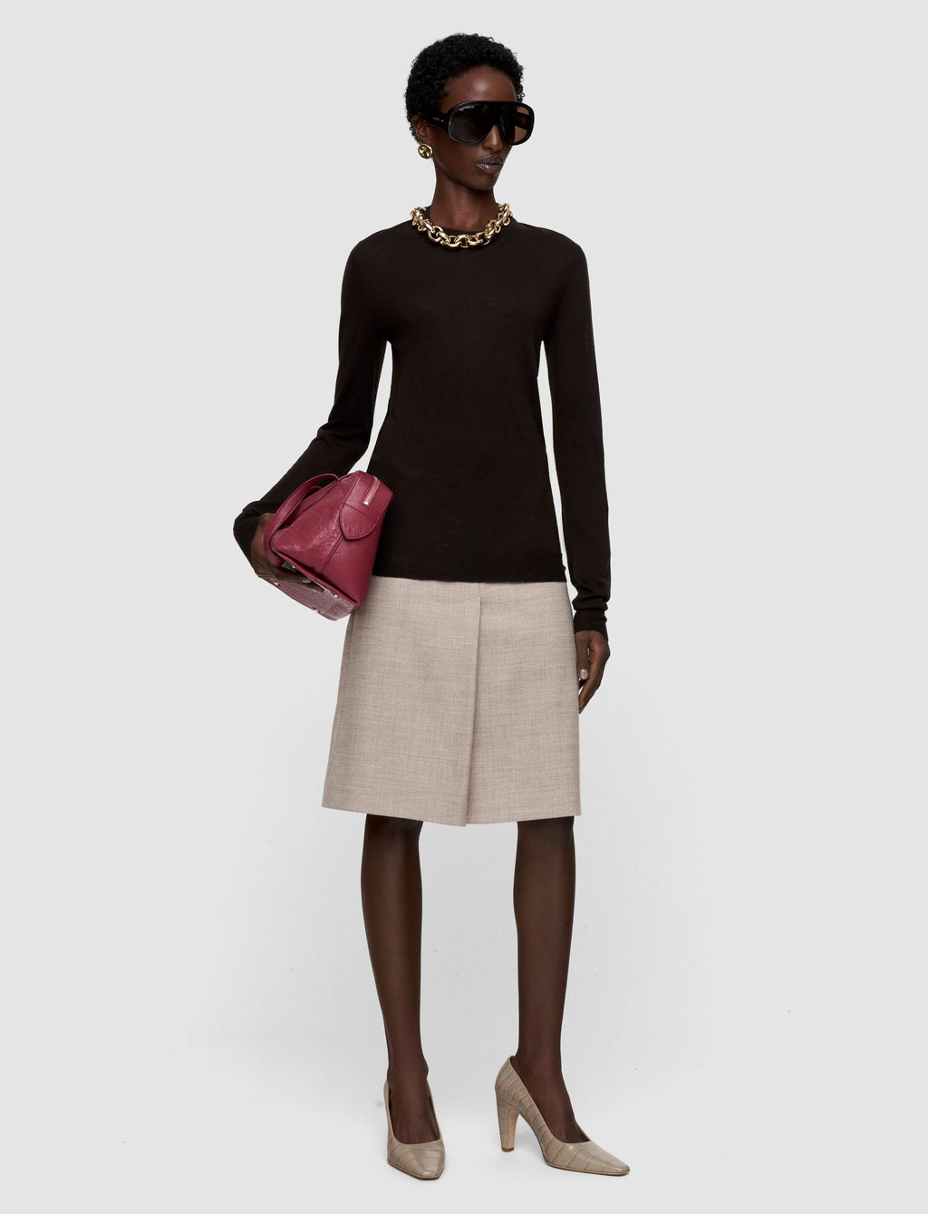 Woman wearing a sheer cocoa brown  long-sleeve top, beige skirt, and holding a pink handbag on a white background