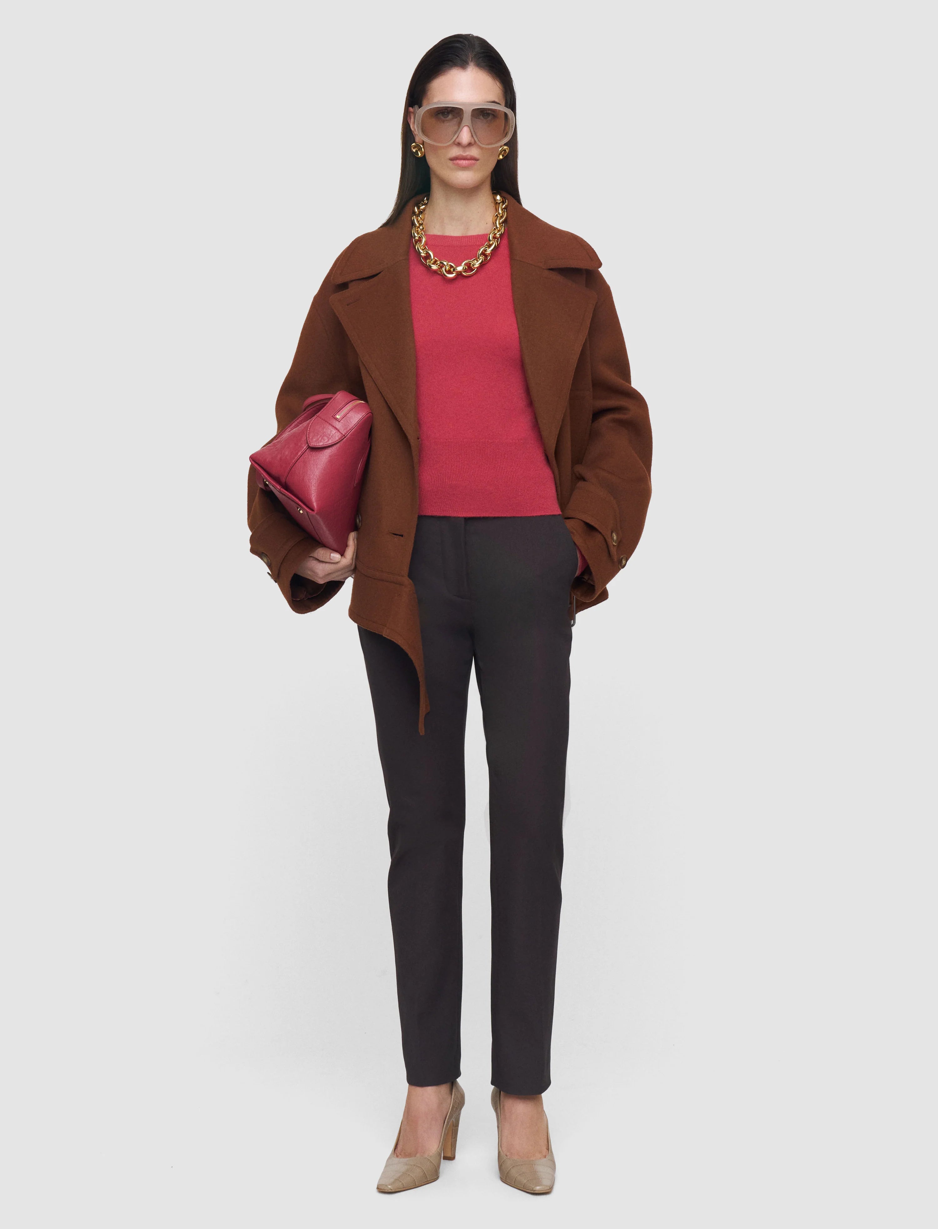Woman wearing a brown coat, red top, black pants, and holding a pink bag on a white background
