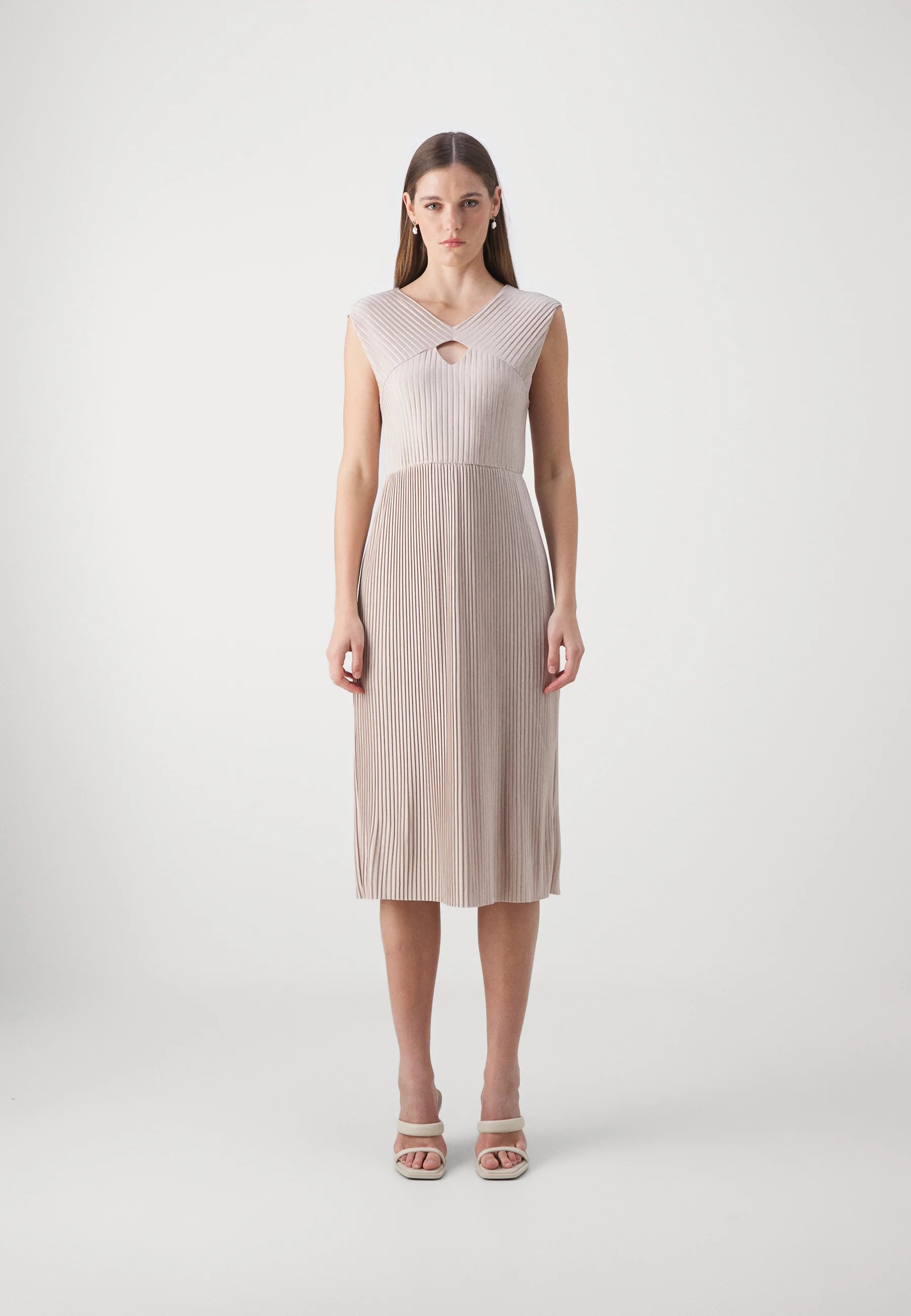 A female model wearing a sleeveless, high-neck, pleated midi dress in a beige color, with a cut-out detail at the neckline.