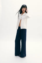 A model stands against a seamless white studio background wearing a white cropped button-up shirt with dark navy wide-leg pants.