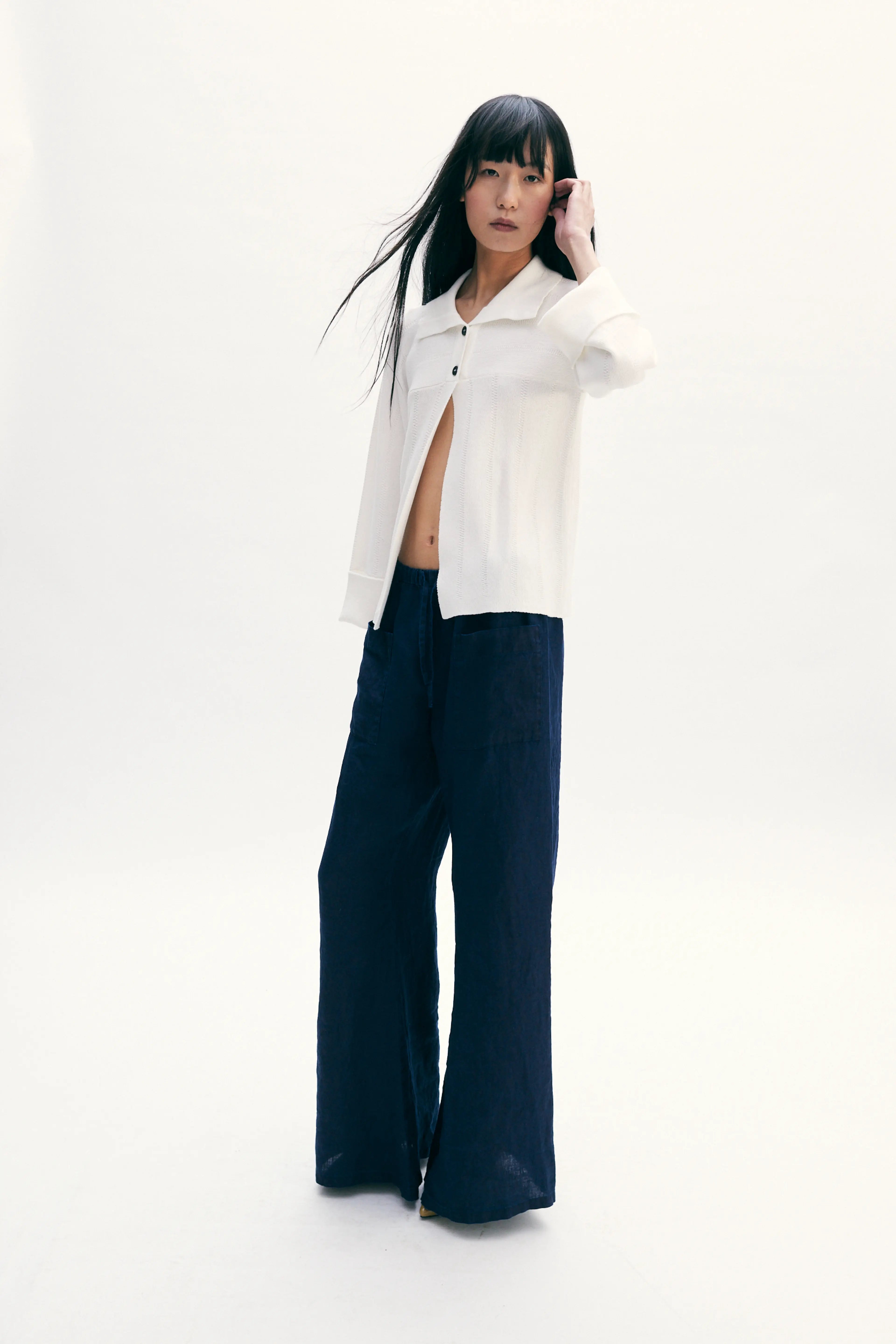 A model stands against a seamless white studio background wearing a white cropped button-up shirt with dark navy wide-leg pants.