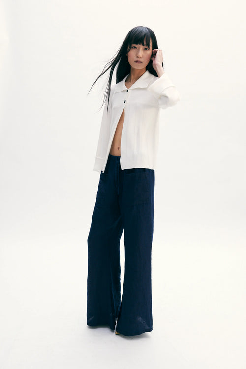 A model stands against a seamless white studio background wearing a white cropped button-up shirt with dark navy wide-leg pants.