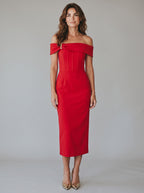 A woman modeling a bright red off-the-shoulder midi dress with structured detailing, paired with pointed-toe heels, against a neutral light gray studio background.