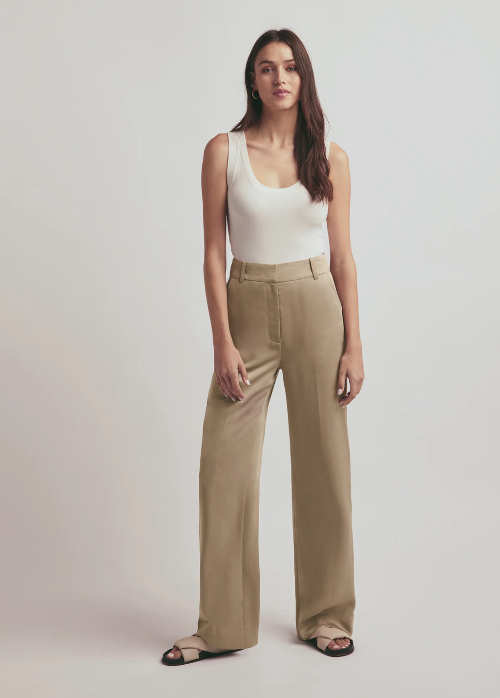 Woman wearing a white tank top and beige pants on a plain background