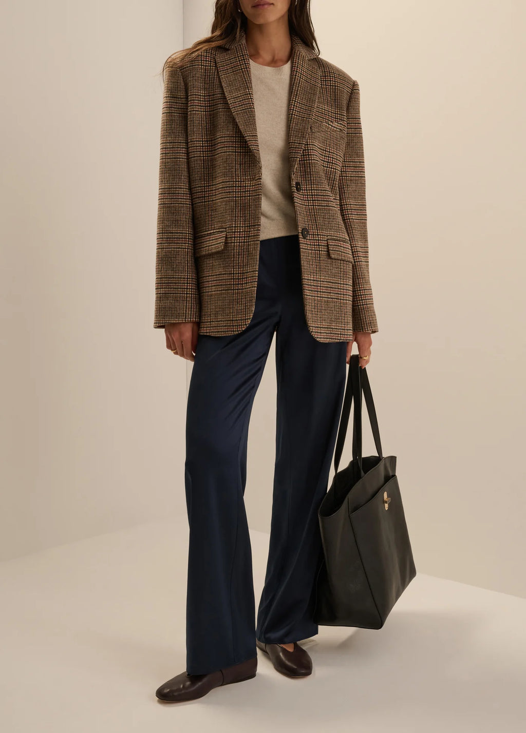 A model stands in a studio wearing an oversized brown plaid blazer over a cream top, navy wide-leg trousers, and brown loafers, while holding a large black tote bag.