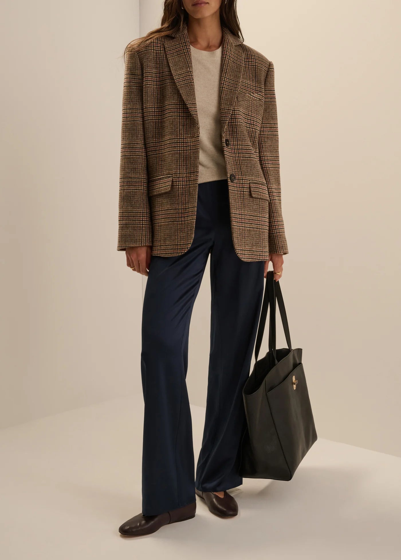 A model stands in a studio wearing an oversized brown plaid blazer over a cream top, navy wide-leg trousers, and brown loafers, while holding a large black tote bag.