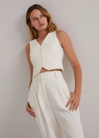 A person wearing a sleeveless, buttoned, ivory-colored vest with a V-neckline, paired with white trousers.
