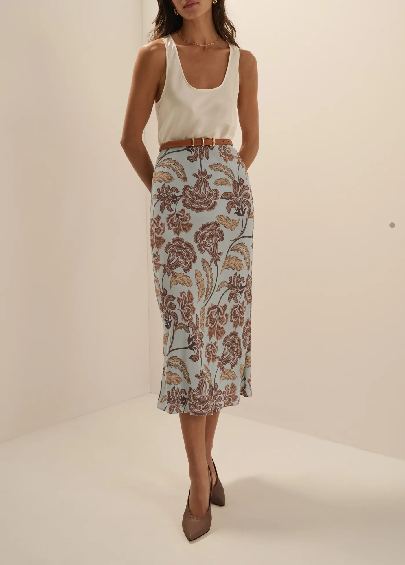 A model wearing a sleeveless white top and a high-waisted midi skirt with a brown and beige floral pattern on a light blue background, styled with a thin brown belt and brown pointed-toe heels.