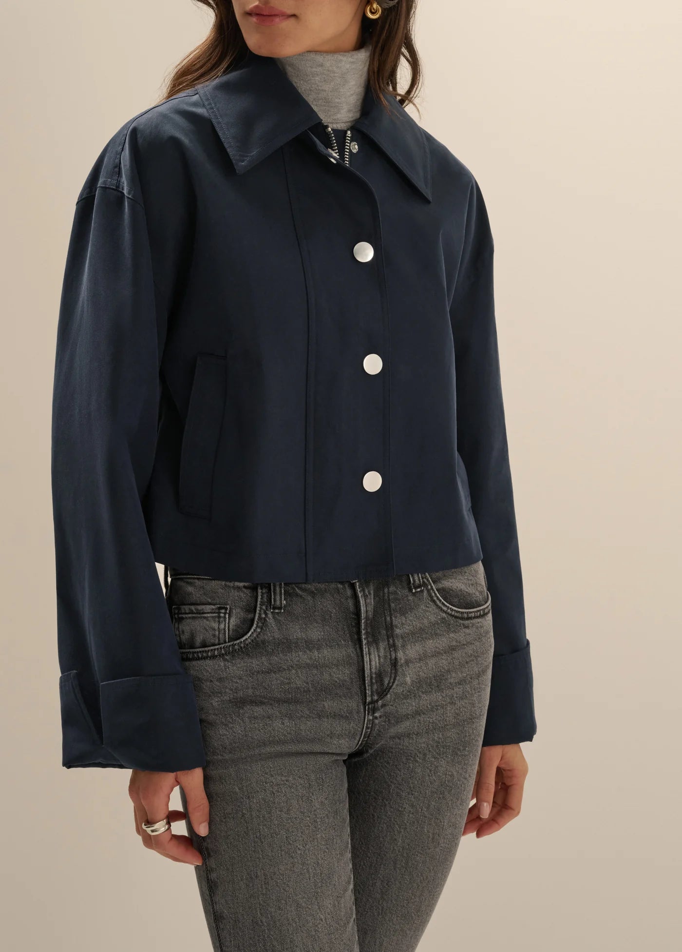 A woman wearing a cropped navy blue jacket with white buttons over a gray turtleneck and gray jeans, shown from the neck down against a plain beige background.