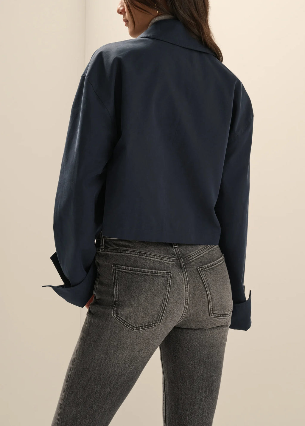 A woman shown from the back wearing a dark navy cropped button-up shirt and gray jeans against a plain beige studio background.