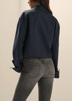 A woman shown from the back wearing a dark navy cropped button-up shirt and gray jeans against a plain beige studio background.