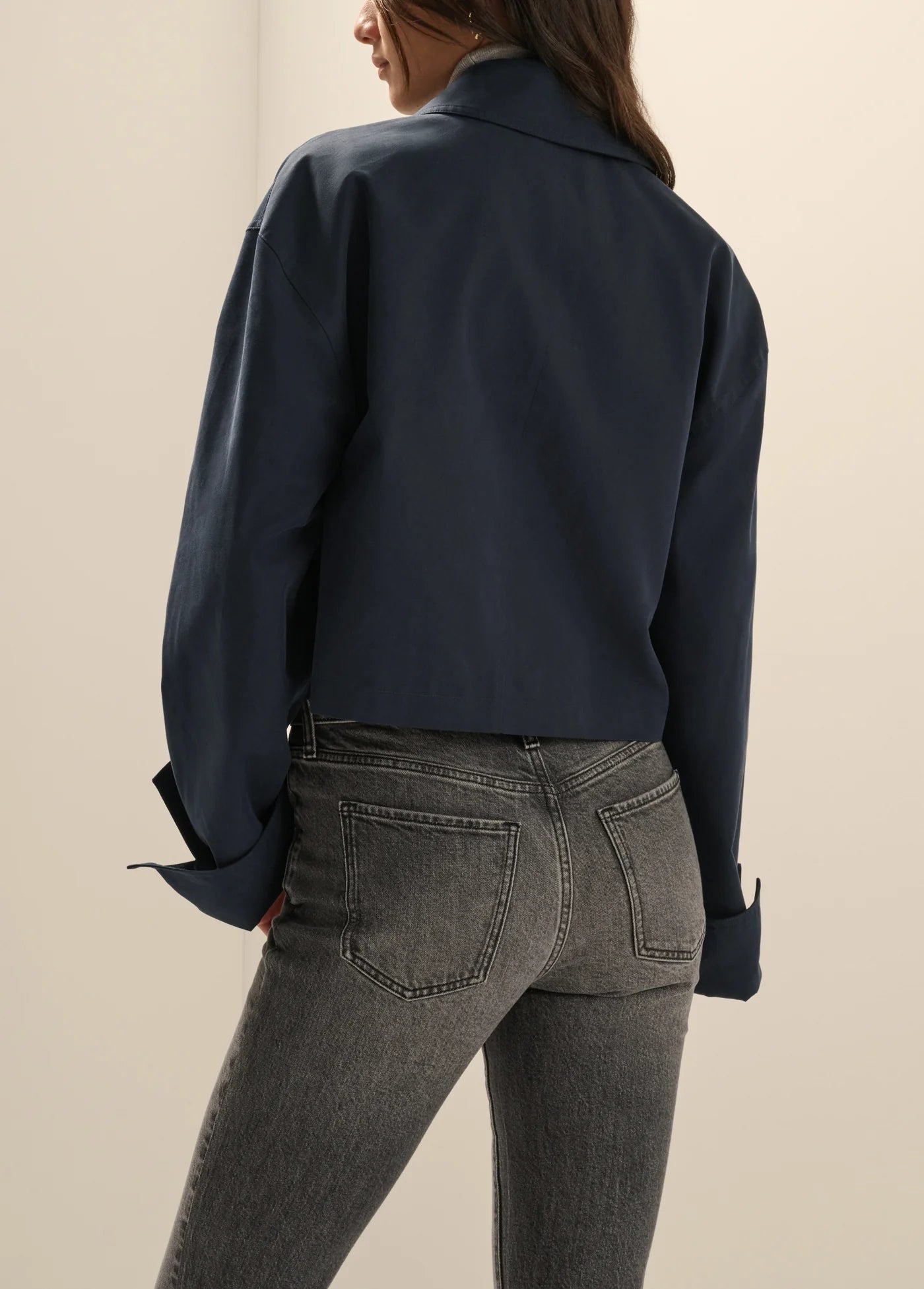 A woman shown from the back wearing a dark navy cropped button-up shirt and gray jeans against a plain beige studio background.