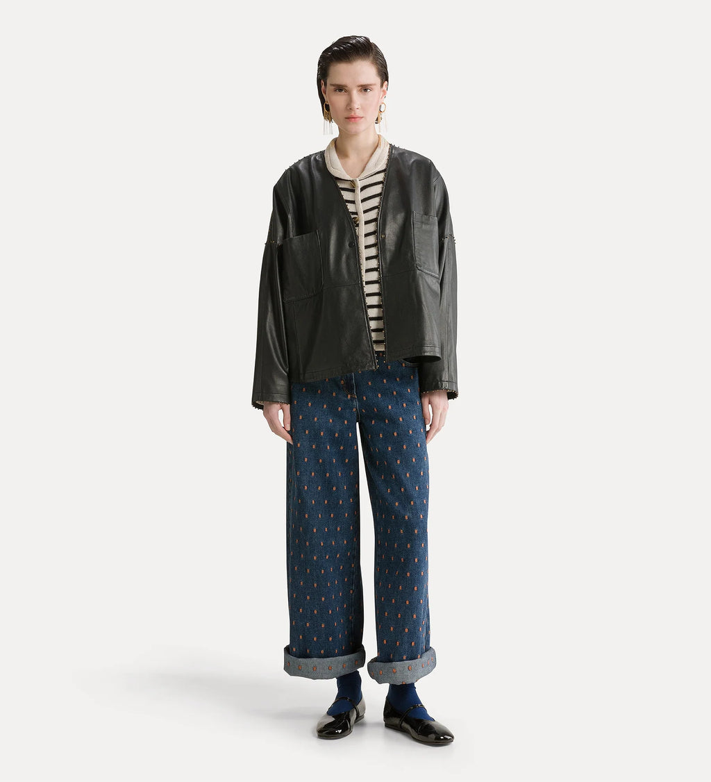 A model stands against a light gray studio background wearing a black leather jacket over a cream and black striped shirt, paired with wide-leg blue patterned trousers, dark socks, and black shoes.