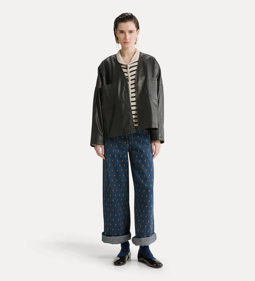 A model stands against a light gray studio background wearing a black leather jacket over a cream and black striped shirt, paired with wide-leg blue patterned trousers, dark socks, and black shoes.