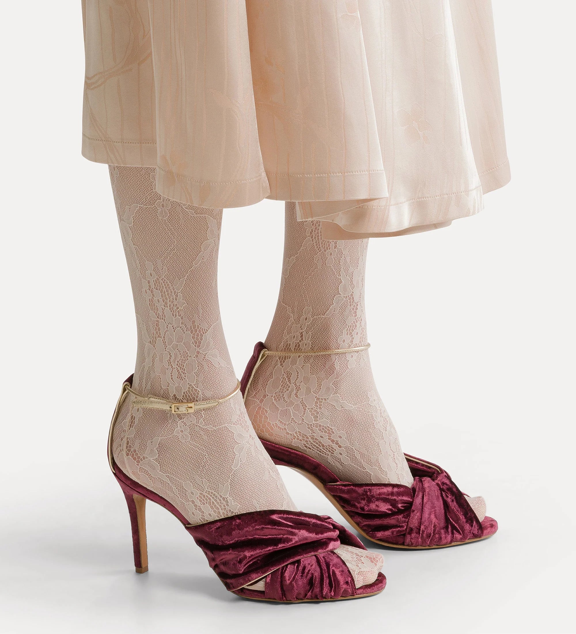 Maroon high-heeled shoes with lace detailing worn with a beige skirt and tights on a white background