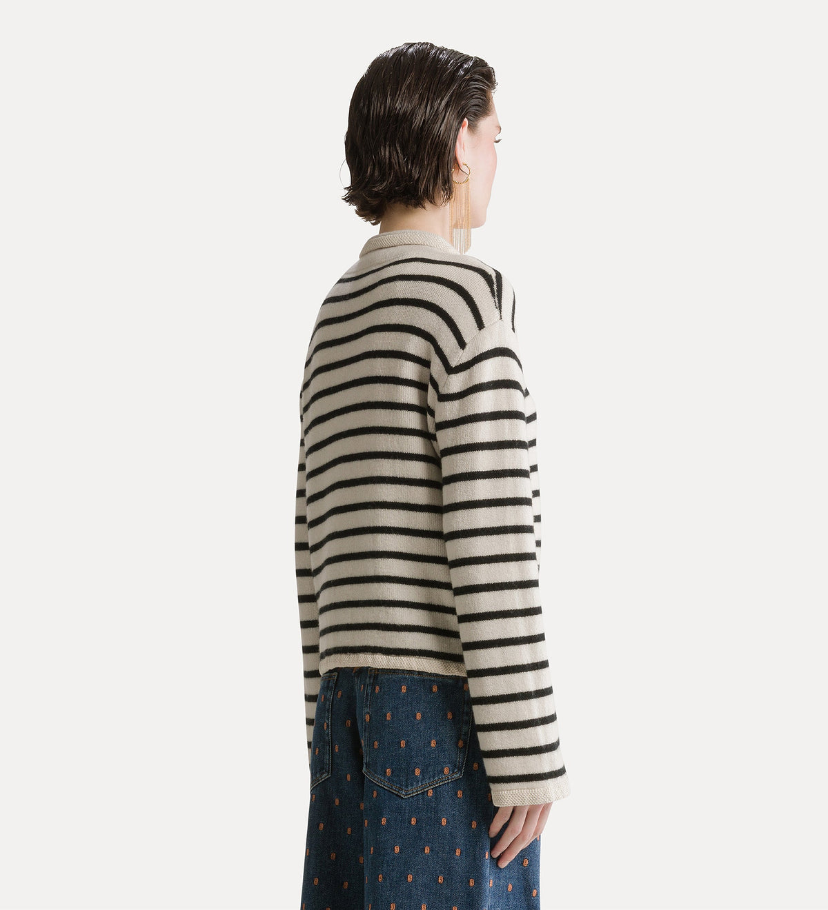 A model shown from the back wearing a cream-colored knit sweater with thin black horizontal stripes and a high collar, paired with a blue patterned skirt, against a solid off-white background.