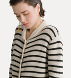 A close-up of a woman wearing a cream-colored knit cardigan with black horizontal stripes and a thick cream rope tie closure, set against a plain light background.
