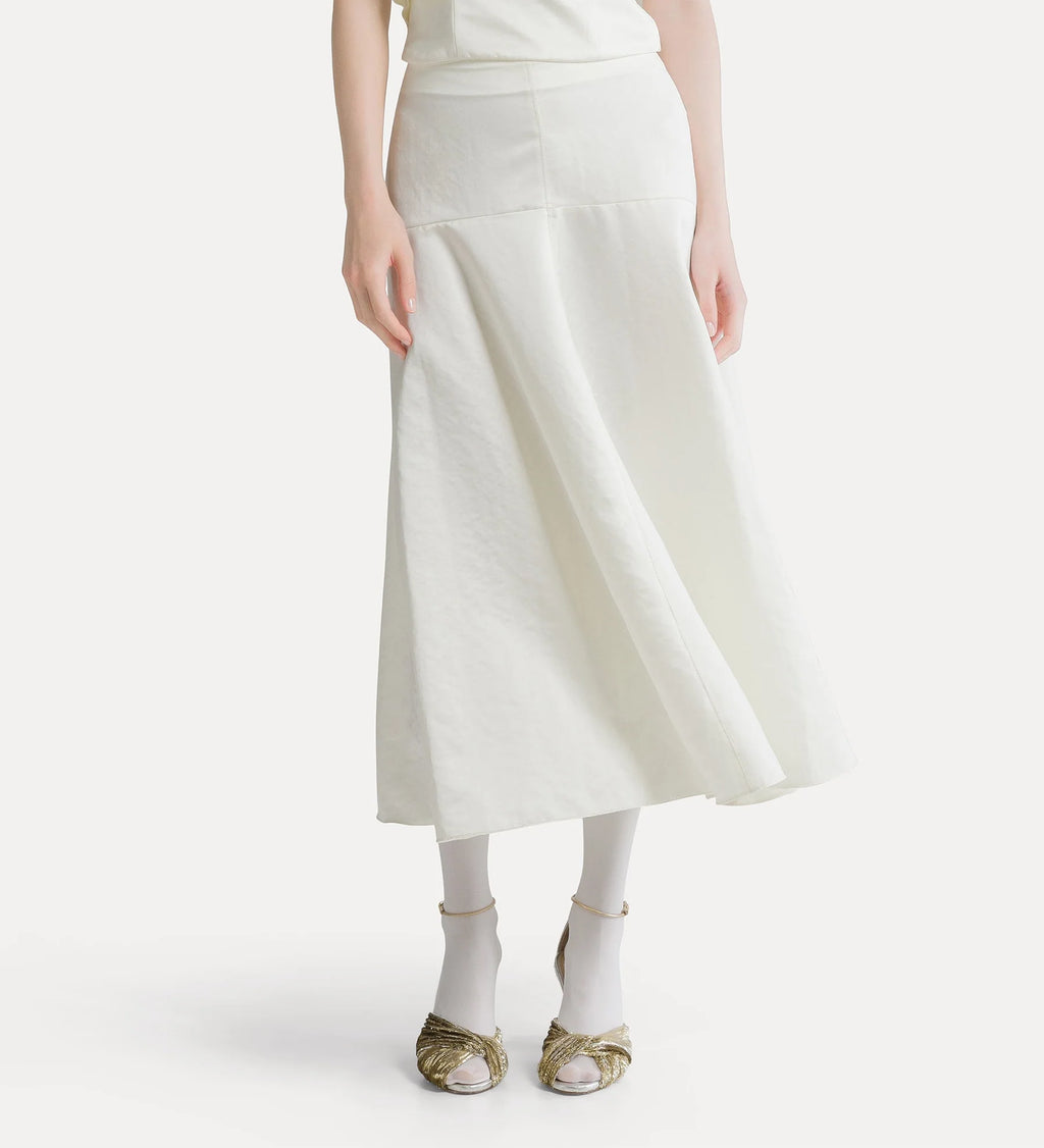 A model wearing a cream-colored linen midi skirt with a smocked waistband, paired with white socks and metallic gold sandals, against a plain off-white background.