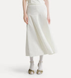 A model wearing a cream-colored linen midi skirt with a smocked waistband, paired with white socks and metallic gold sandals, against a plain off-white background.