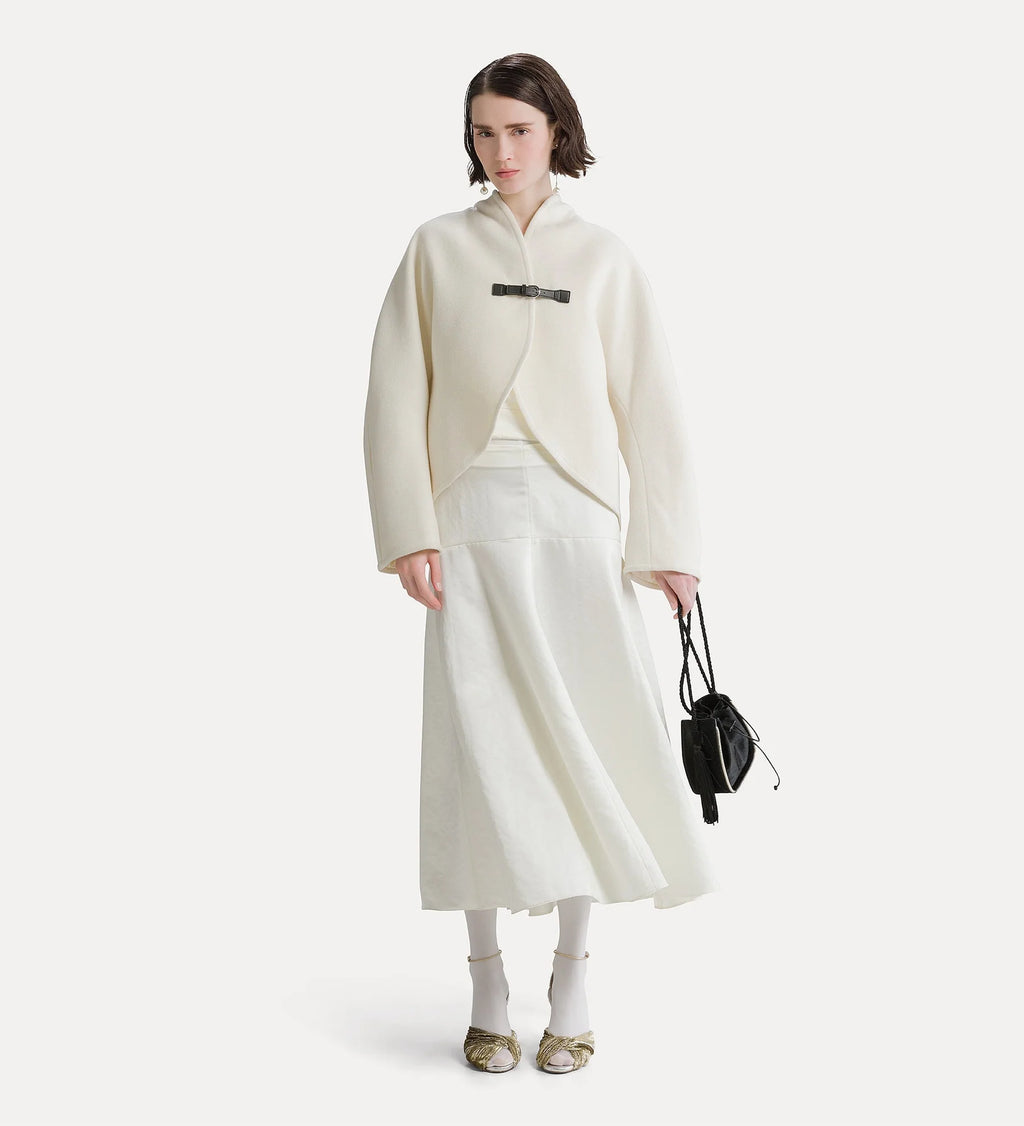 A model stands against a light gray studio background wearing a cream-colored wrap jacket with a black belt detail, a matching cream midi skirt, white socks, and gold flats, while holding a small black handbag.