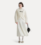 A model stands against a light gray studio background wearing a cream-colored wrap jacket with a black belt detail, a matching cream midi skirt, white socks, and gold flats, while holding a small black handbag.