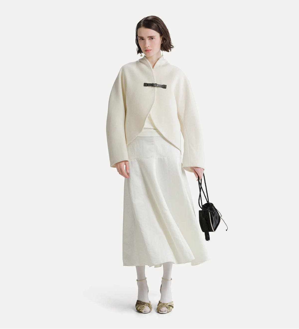 A model stands against a light gray studio background wearing a cream-colored wrap jacket with a black belt detail, a matching cream midi skirt, white socks, and gold flats, while holding a small black handbag.
