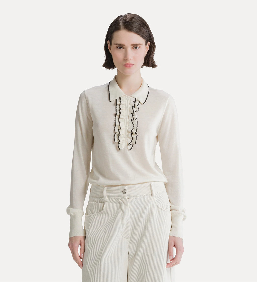 A model wearing a cream-colored long-sleeve knit top with black embroidered ruffles and a pointed collar, paired with cream trousers, against a light gray studio background.