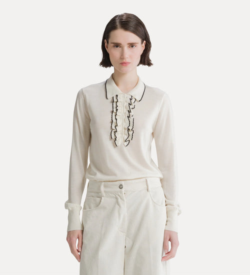 A model wearing a cream-colored long-sleeve knit top with black embroidered ruffles and a pointed collar, paired with cream trousers, against a light gray studio background.